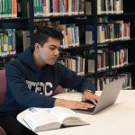 Estudante usa um laptop e consulta livros durante uma sessão de estudos em uma biblioteca universitária.