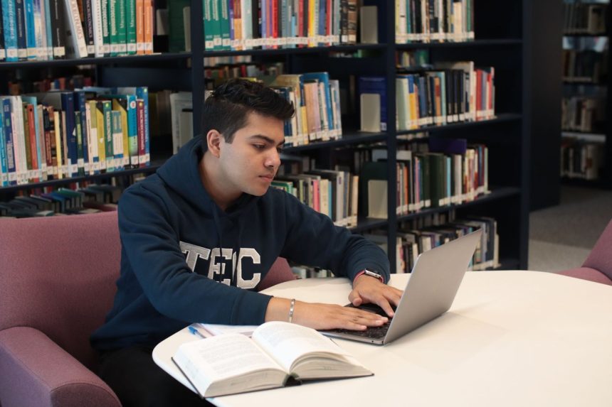 Estudante usa um laptop e consulta livros durante uma sessão de estudos em uma biblioteca universitária.
