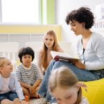 Professora lê um livro para crianças da pré-escola em uma sala de aula colorida.