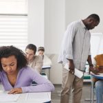 Grupo diverso de estudantes faz uma prova em sala de aula enquanto um professor supervisiona.