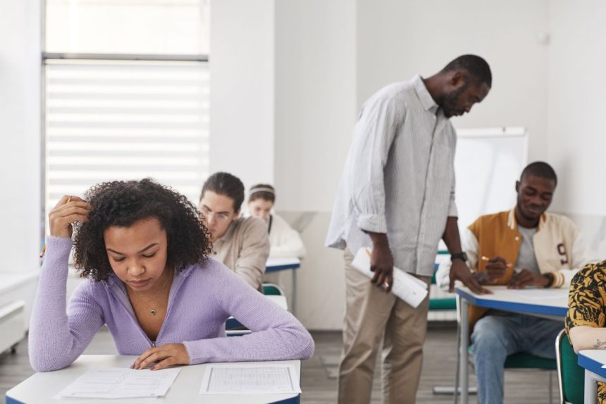 Grupo diverso de estudantes faz uma prova em sala de aula enquanto um professor supervisiona.