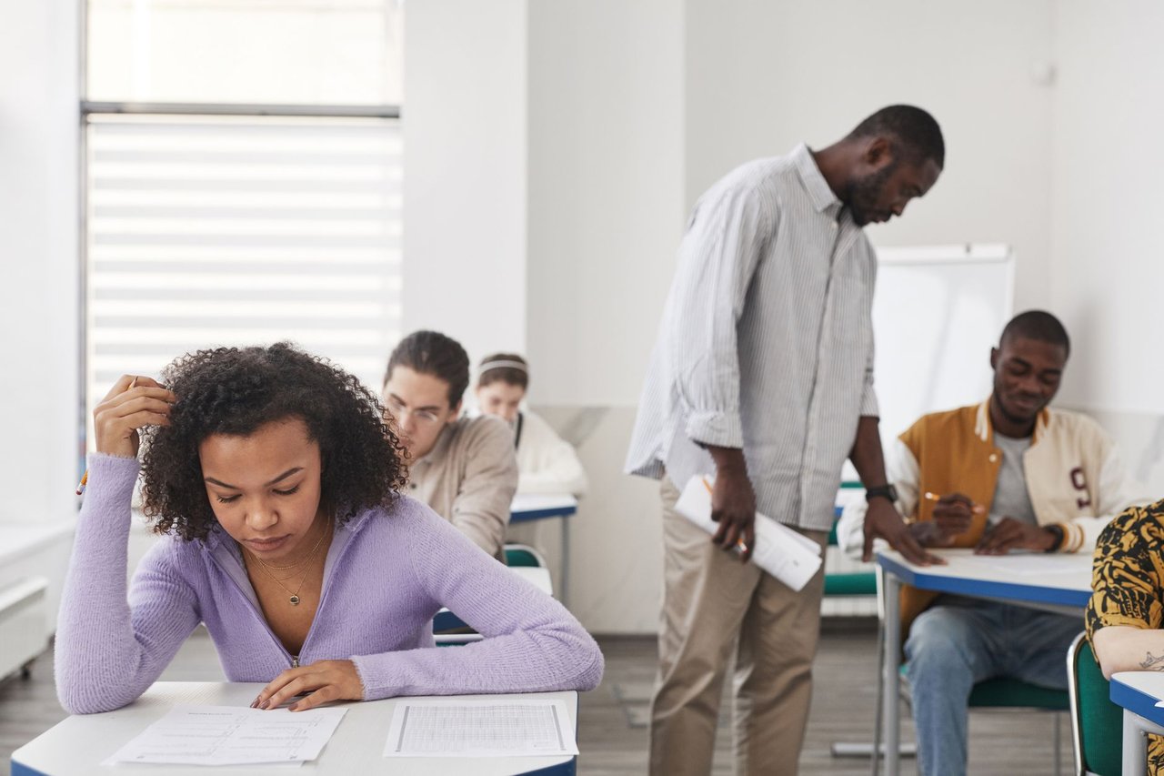 Grupo diverso de estudantes faz uma prova em sala de aula enquanto um professor supervisiona.